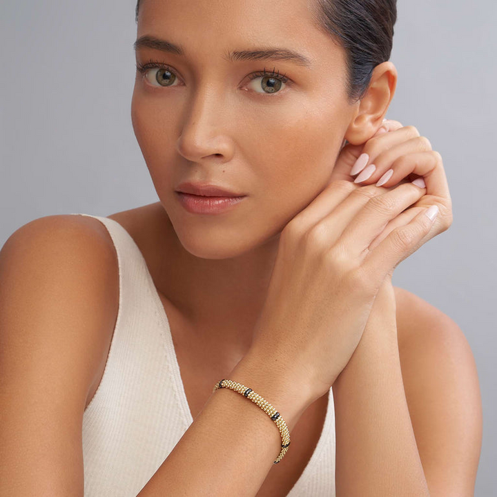 A close-up of a woman wearing a white blouse and an 18k gold bracelet. The bracelet features smooth black stations and caviar beading.