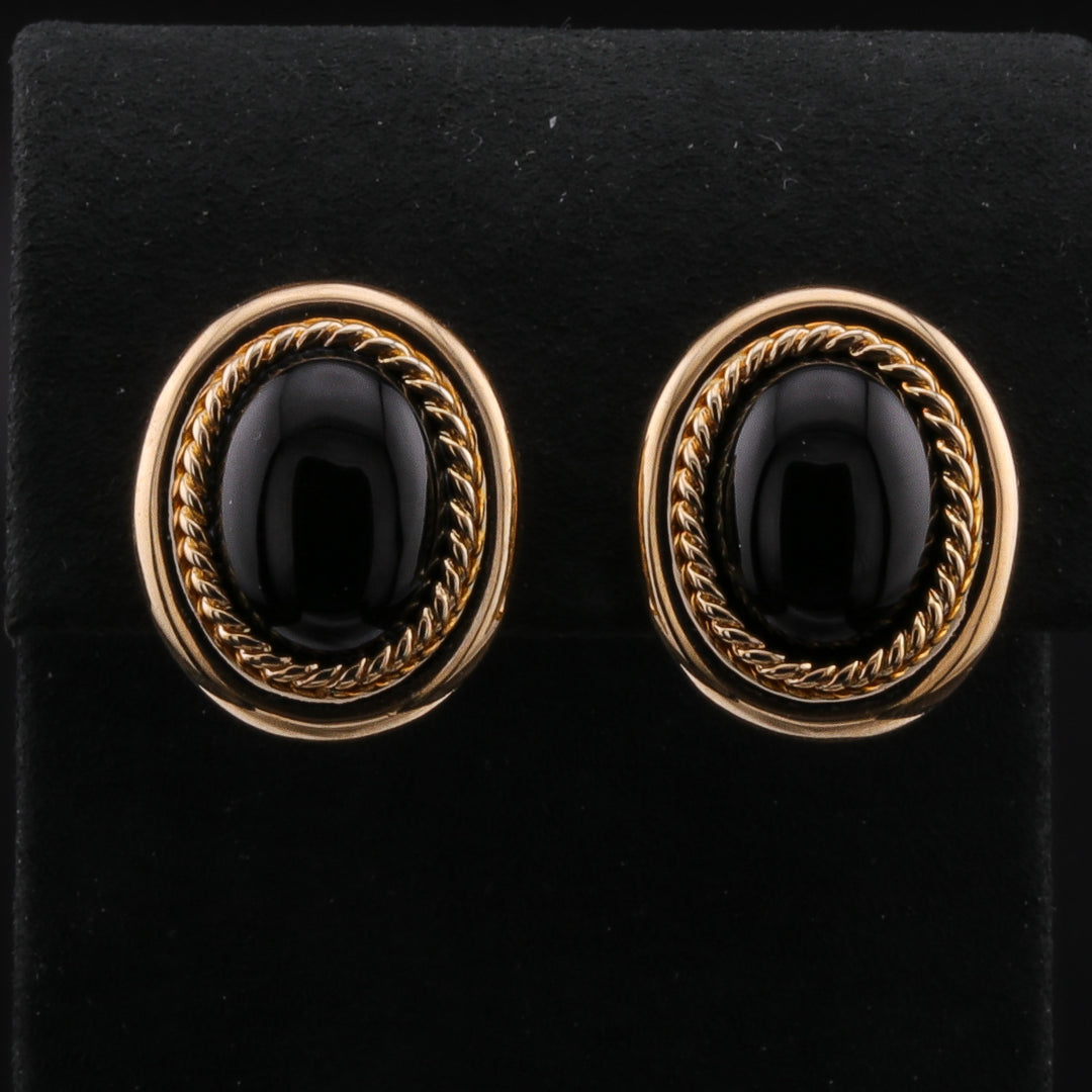 A pair of clip-on onyx earrings with yellow gold cabochon border and deep black onyx stones with a black background.