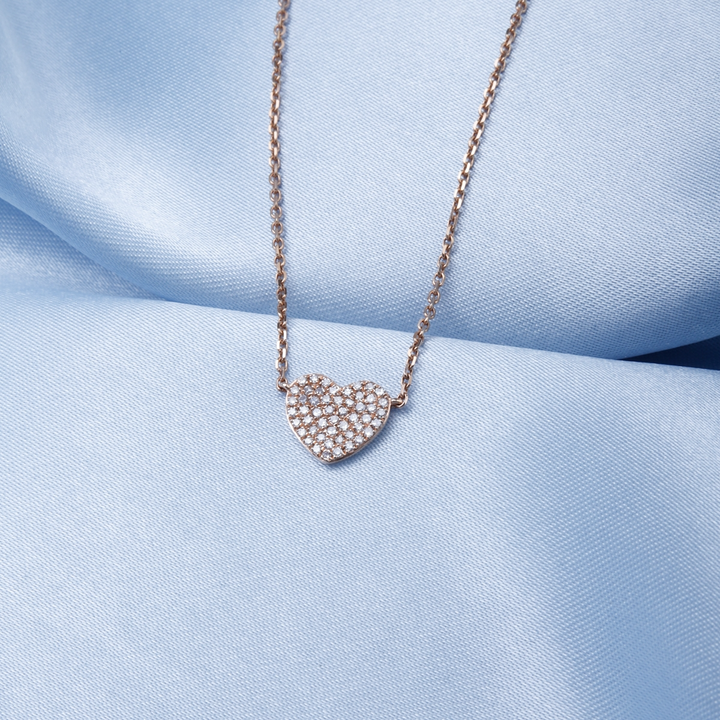 A close-up of a yellow-gold Pave Heart Necklace against a blue background. The necklace features a heart-shaped pendant with round diamonds.