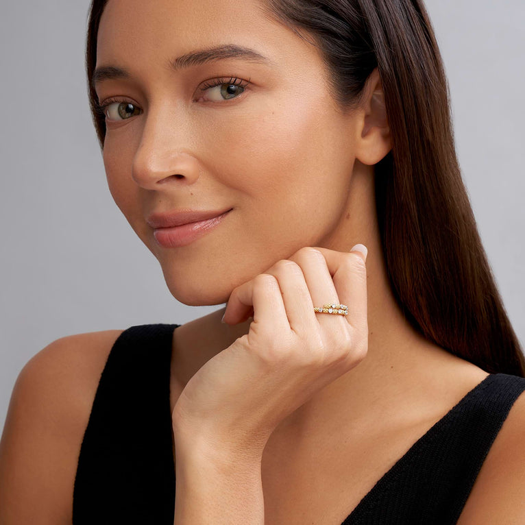 A close-up of a woman with long brown hair, a black tank top, and wearing a ring on her middle finger. The ring features 18k gold superfine Caviar beading, wrap design, and round diamond stations.