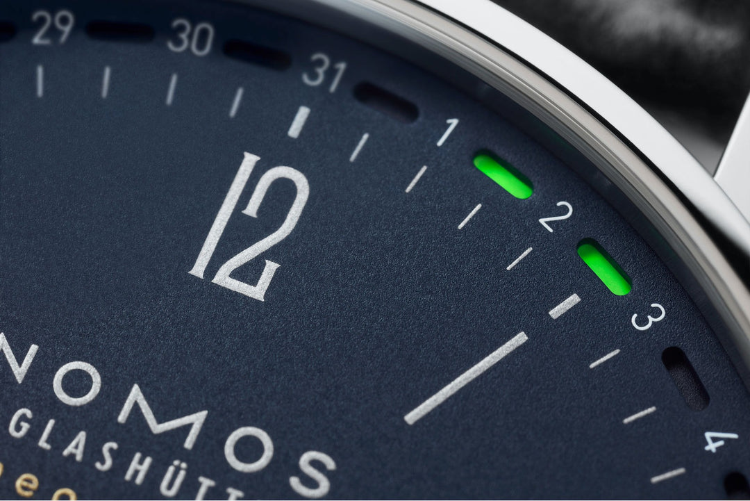 This is a close-up image of a Nomos Glashutte watch angled to the right, focusing on the top right part of the dial. It features a blue dial, silver hands and markers, and a stainless steel bezel.