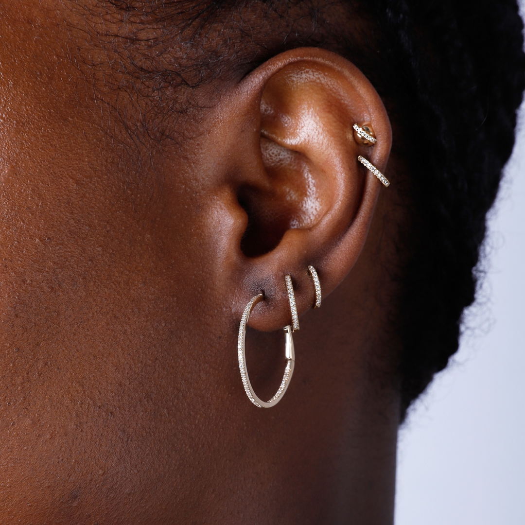 A woman with short black hair is wearing a pair of hoop earrings with a single row of round diamonds.