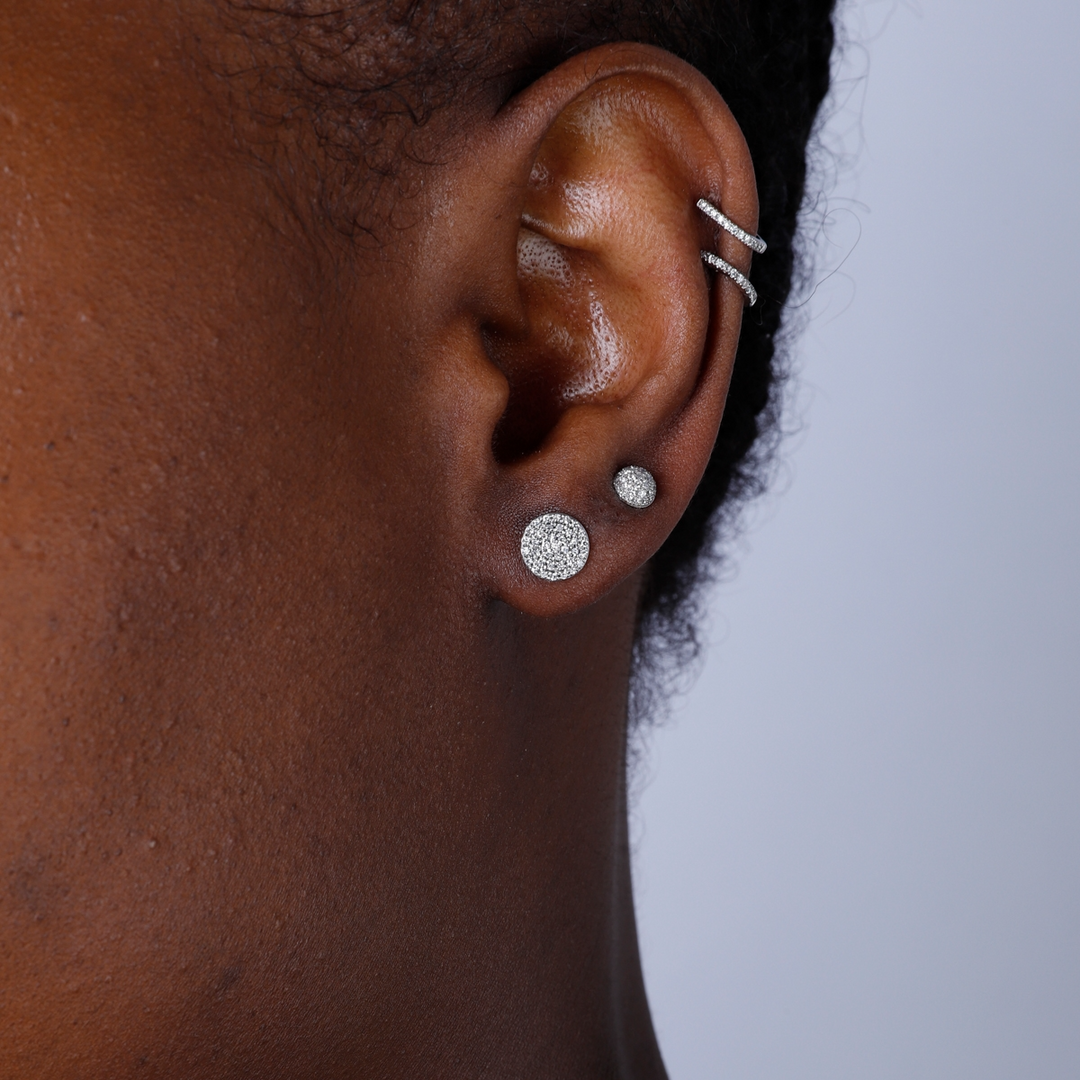 A woman with short black hair is wearing a pair of stud earrings with round, brilliant-cut diamonds framed in a pave setting. 