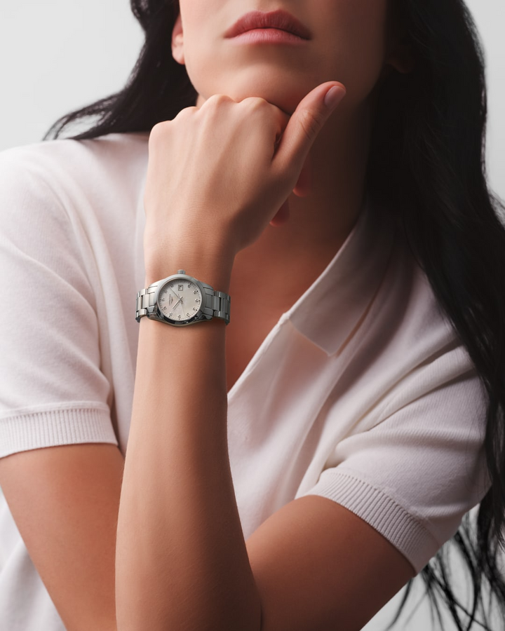 A woman wearing a pink collared top and a Longines watch around her wrist. The watch features a white mother-of-pearl diamond-set dial, silver hands and markers, a stainless steel bezel, and a stainless steel bracelet.