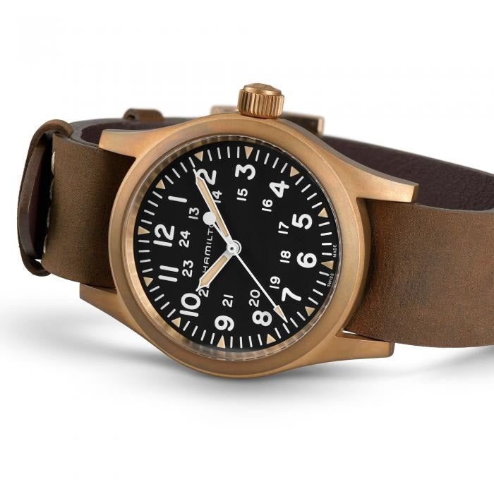 A Hamilton watch is lying on its left side against a white background. It features a black dial, white hands and markers, a bronze case, a bronze bezel, a bronze crown, and a brown nato leather strap.