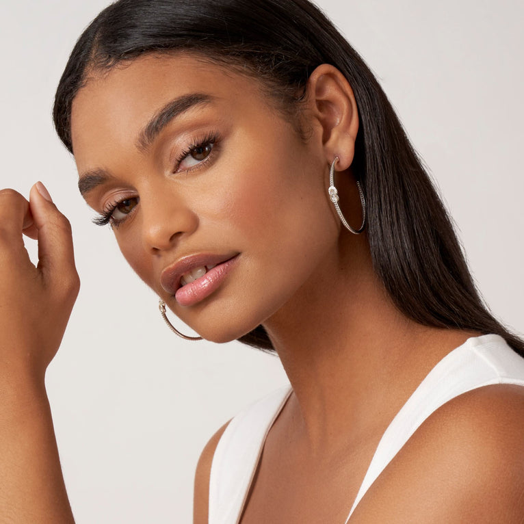 A woman is wearing a white tank top and a pair of hoop earrings with diamonds and 18K gold woven in a knot motif. 