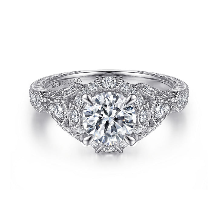 A white gold ring lies flat against a white background. It features a round-cut diamond set in the center with a diamond halo and petal-shaped diamond clusters.