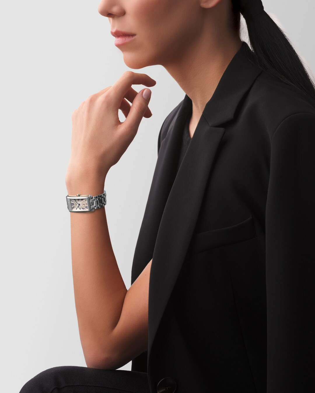 A woman wearing a black blouse and a Longines watch around her wrist. The rectangular-shaped watch features a white dial, blue hands, black markers, a stainless steel bezel, and a stainless steel bracelet.