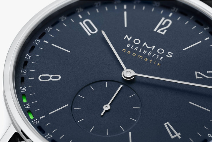 This is a close-up image of a Nomos Glashutte watch angled to the right, focusing on the dial. It features a blue dial, silver hands and markers, a stainless steel bezel, and a black leather strap.