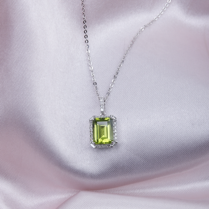 A close-up of a white-gold Delicate Emerald Cut Necklace against a pink background. The necklace features a cushion-cut light green center stone and an 18k white gold chain.