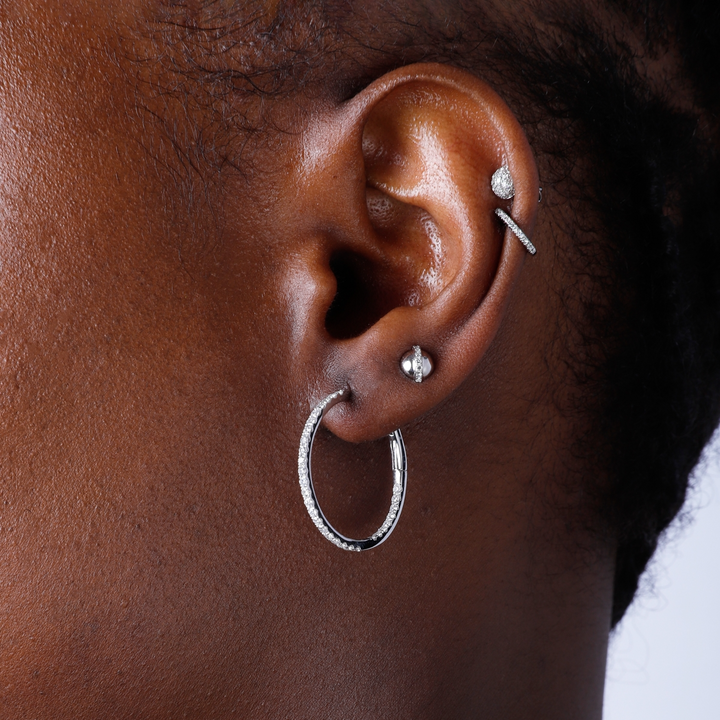 A woman with short black hair is wearing a pair of white gold hoop earrings with a single row of round diamonds.