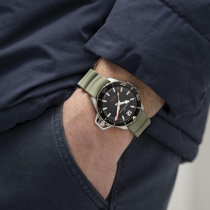 A man is wearing a Hamilton watch. It features a black dial, white hands and markers, a steel case, a black bezel, a steel crown, and a green rubber strap.