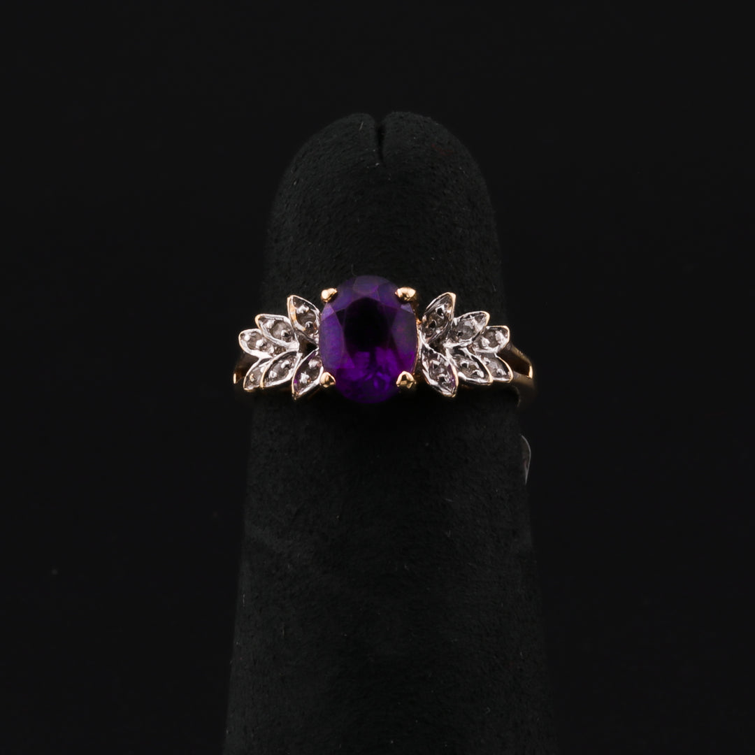 This is a close-up of a yellow-gold ring with an amethyst center stone and marquise diamond side stones artfully set to resemble delicate leaves. The ring rests on a black ring holder against a black background.