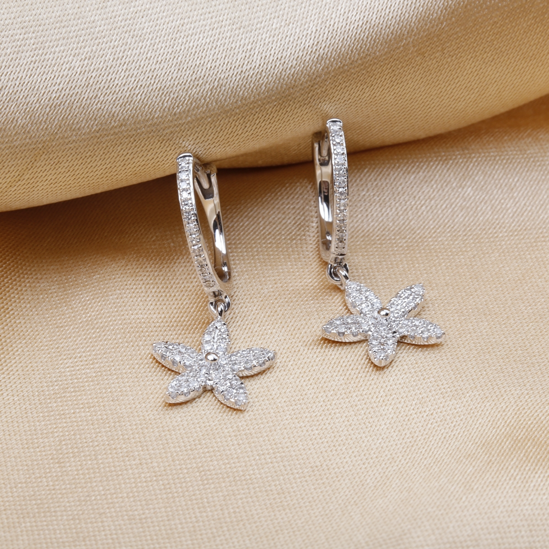 A pair of drop diamond earrings. The earrings have a flower design and a cluster of diamonds against a gold background.