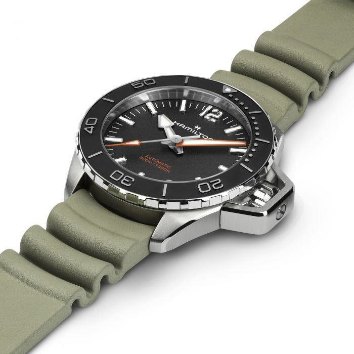 A Hamilton watch is laid out tilted to the right against a white background. It features a black dial, white hands and markers, a steel case, a black bezel, a steel crown, and a green rubber strap.