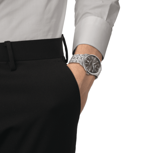 A man wearing a button-down and black pants with a Tissot watch on his wrist. The watch features a gray dial, steel bezel, silver hands and markers, and a stainless steel bracelet.