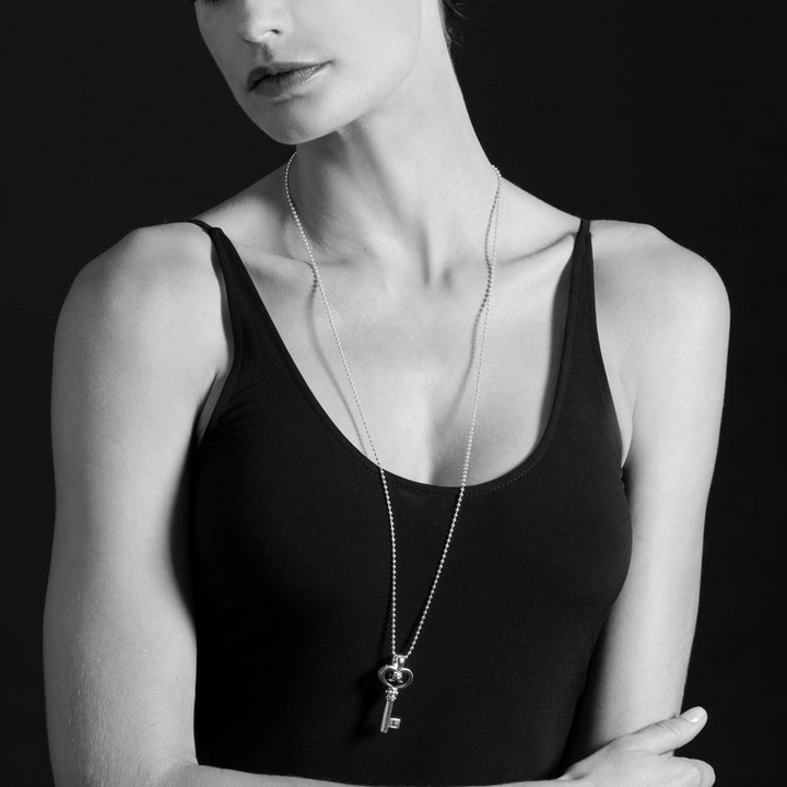 A woman is wearing a sterling silver necklace in the middle of the white background featuring a key pendant with Caviar beading, fluted accents, the LAGOS logo, and crest finished on a ball chain necklace