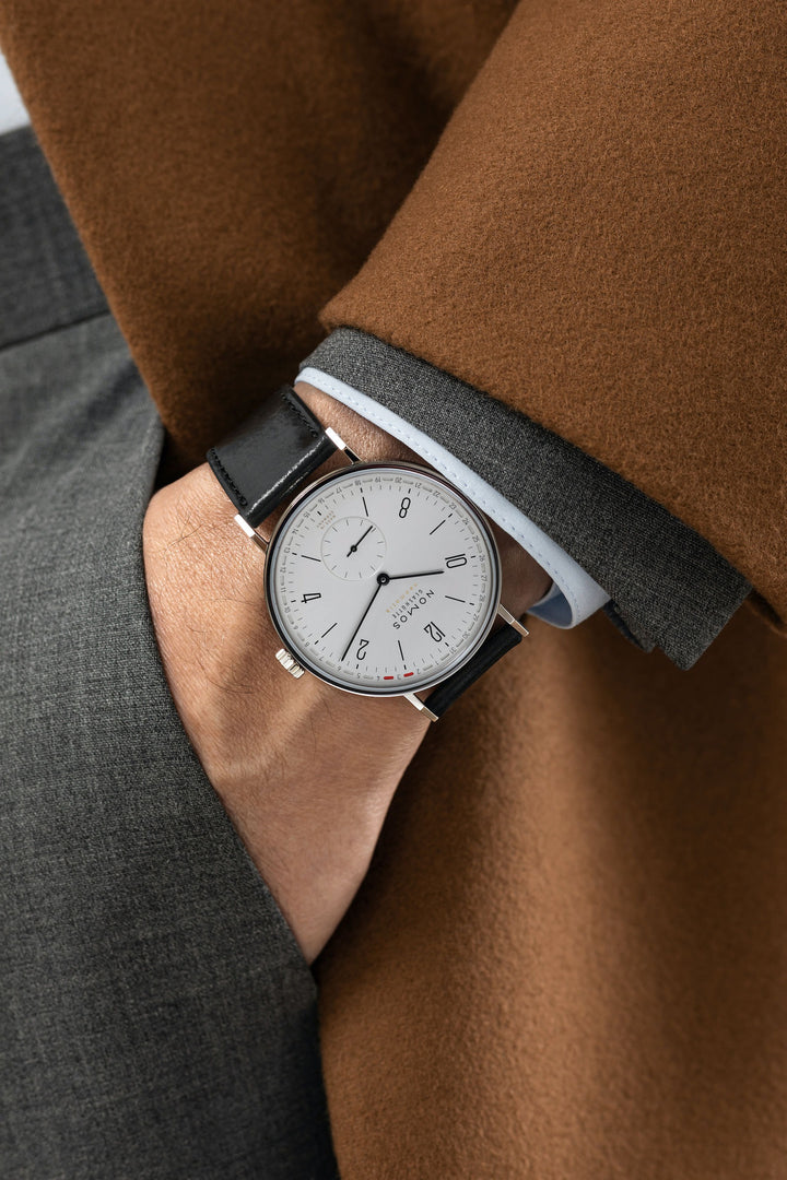 A man wearing a Nomos Glashutte watch on his wrist. It features a white dial, black hands and markers, a stainless steel bezel, and a black leather strap.