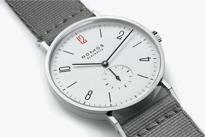 This is a close-up image of a Nomos Glashutte watch angled to the left. It features a white dial, black hands and markers, a stainless steel bezel, and a gray strap.