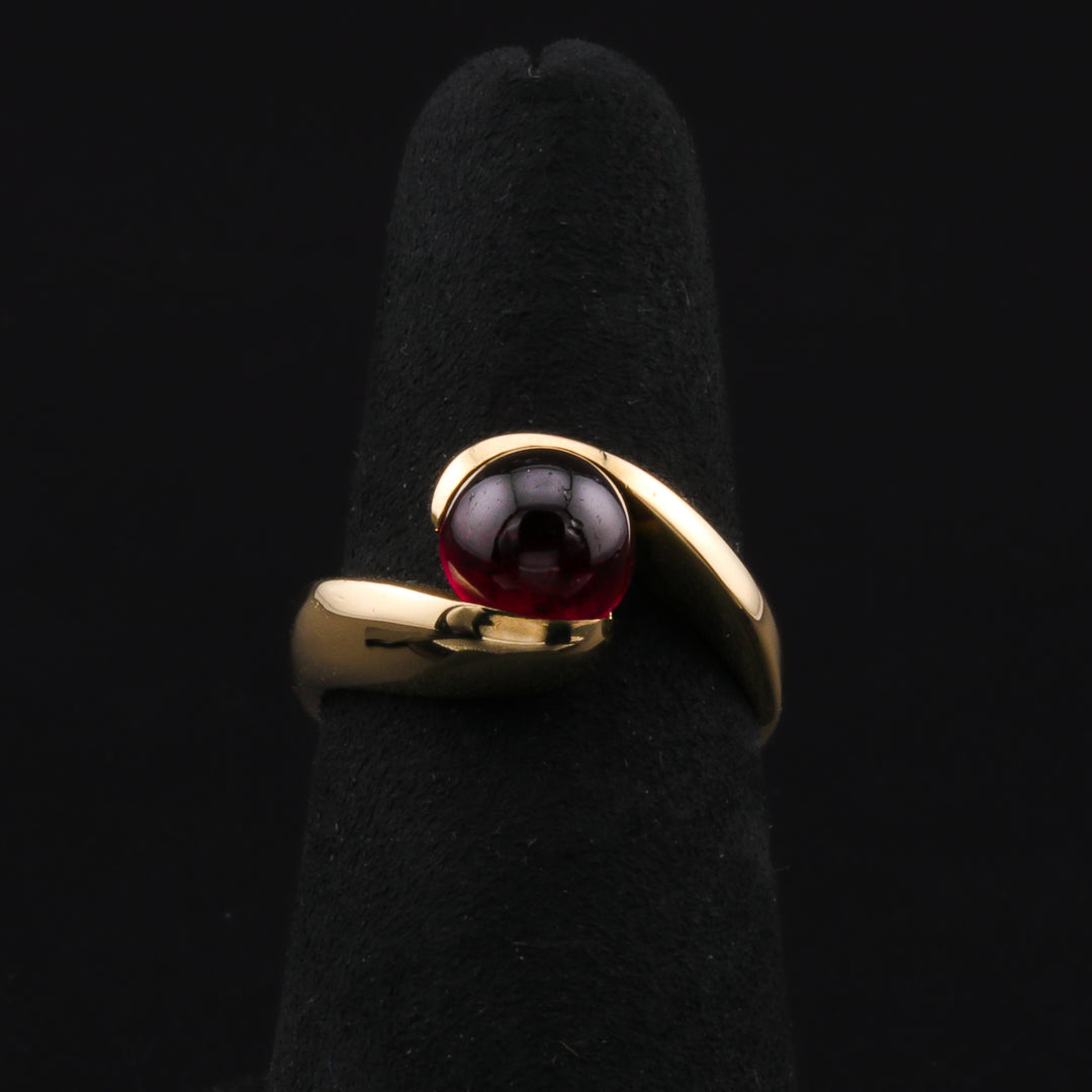 This is a close-up of a yellow-gold ring with a smooth, rounded surface of the cabochon garnet held in place by the tension of the metal. The ring is resting on a black ring holder against a black background.