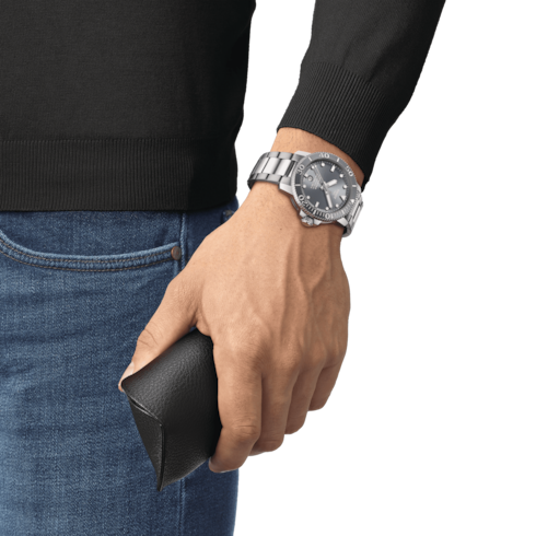 A man wearing a Tissot watch around his wrist.  The watch features a gray dial, a gray bezel, white hands and markers, and a stainless steel bracelet.