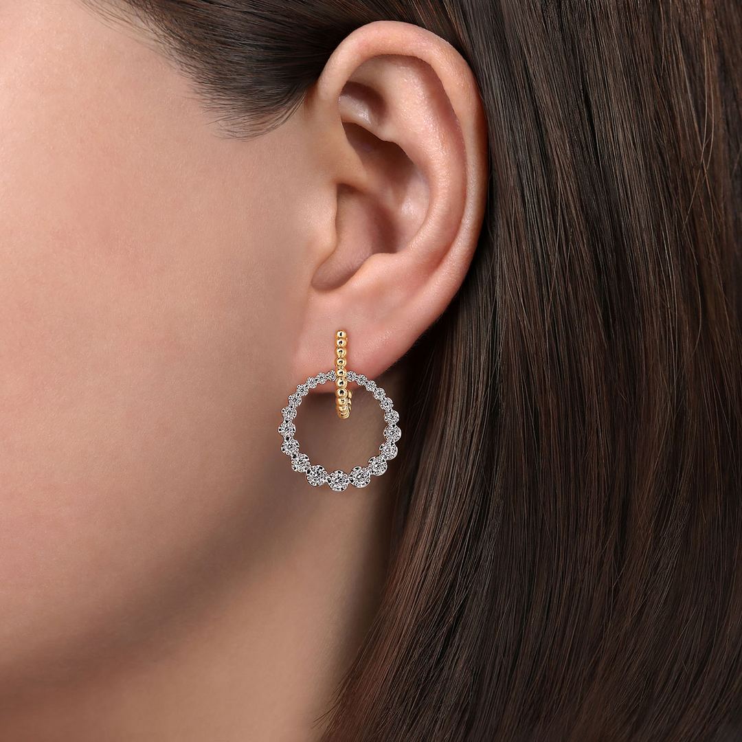 This is a close-up of a woman's ear wearing huggie drop earrings with a circular motif, which combines 14K yellow gold and diamonds.