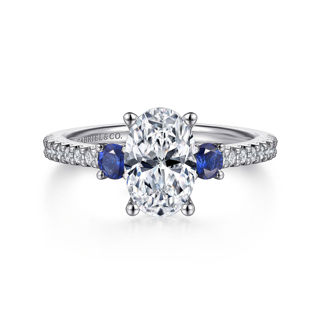 A white gold ring lies flat against a white background. It features a brilliant-cut diamond set in the center, sapphire side stones, and a diamond-set band.