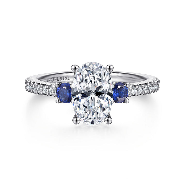 A white gold ring lies flat against a white background. It features a brilliant-cut diamond set in the center, sapphire side stones, and a diamond-set band.