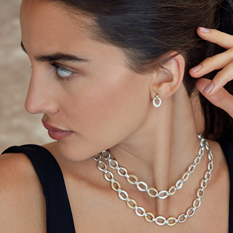 A woman wearing a black tank top, necklace, and a pair of drop earrings with oval drop highlighted by 18k gold Caviar beading. The right earring is angled to the side.