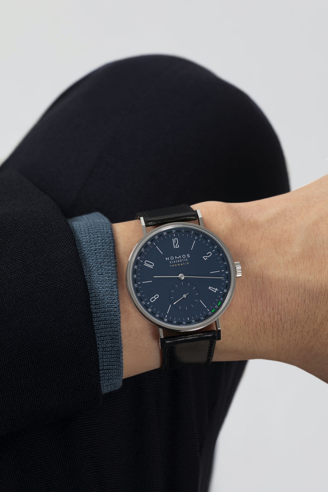 A man wearing a Nomos Glashutte watch on his wrist. It features a blue dial, silver hands and markers, a stainless steel bezel, and a black leather strap.