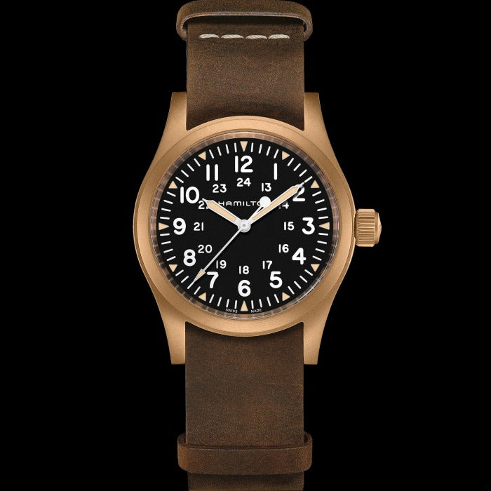 A Hamilton watch is laid out against a black background. It features a black dial, white hands and markers, a bronze case, a bronze bezel, a bronze crown, and a brown nato leather strap.