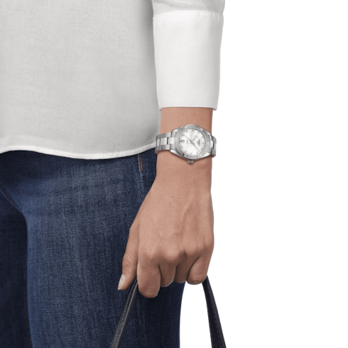 A woman wearing a gray top and jeans with a Tissot watch holding a black purse on her wrist. The watch features a mother-of-pearl diamond-set dial, a steel bezel, silver hands and markers, and a stainless steel bracelet.