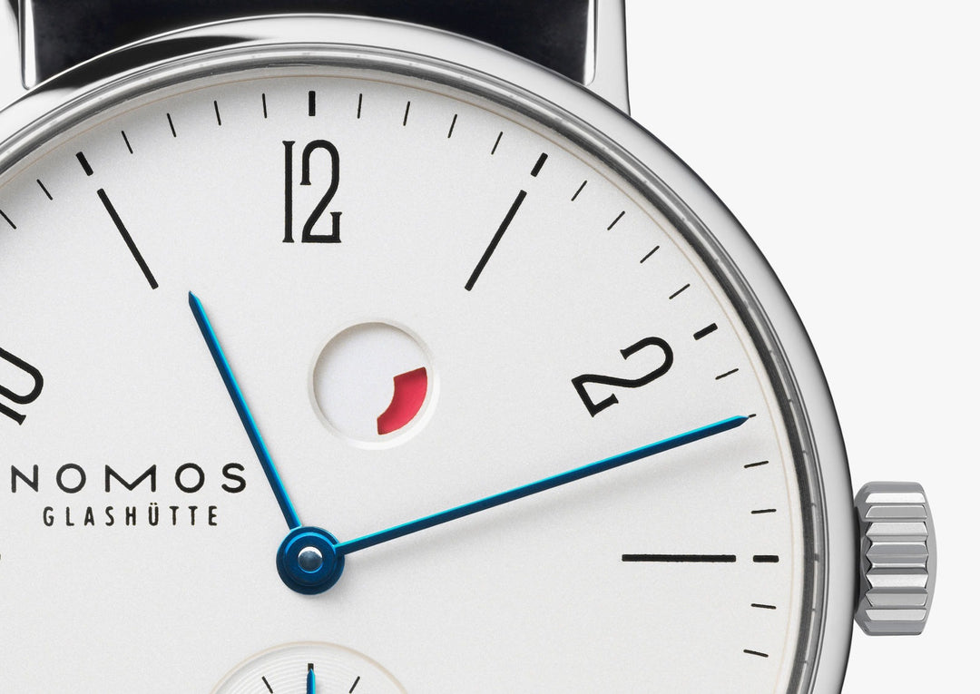 This is a close-up image of a Nomos Glashutte watch, focusing on the top part of the dial. It features a white dial, black & blue hands and markers, a stainless steel bezel, and a black leather strap.