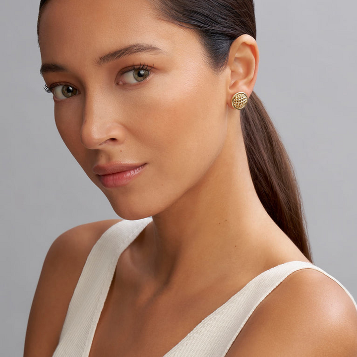 A woman wearing a white tank top and 18k gold Caviar beading is surrounded by gold ceramic detailing on the stud earrings. 