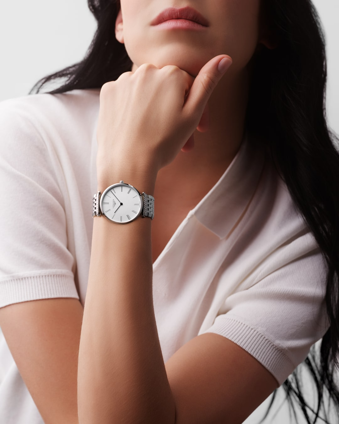 A woman wearing a pink collared shirt and a Longines watch around her wrist. The watch features a white dial, black hands and markers, a stainless steel bezel, and a stainless steel bracelet.