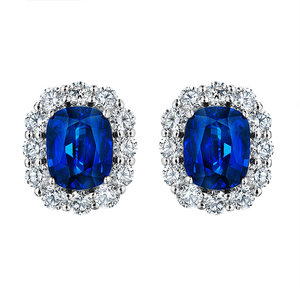 A pair of stud earrings is displayed in the middle of a white background. The earring features a cushion-cut sapphire center stone with surrounding diamonds.