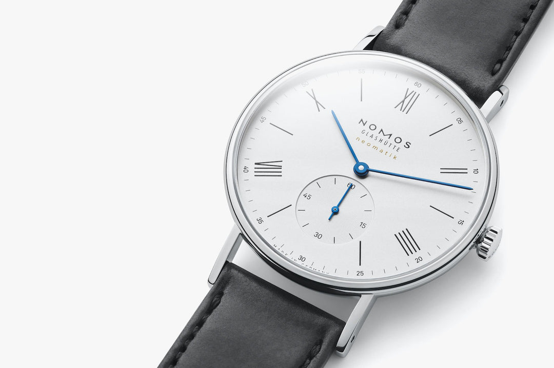 A close-up image of a Nomos Glashutte watch angled to the right. It features a white dial, blue hands and markers, a stainless steel bezel, and a black strap.