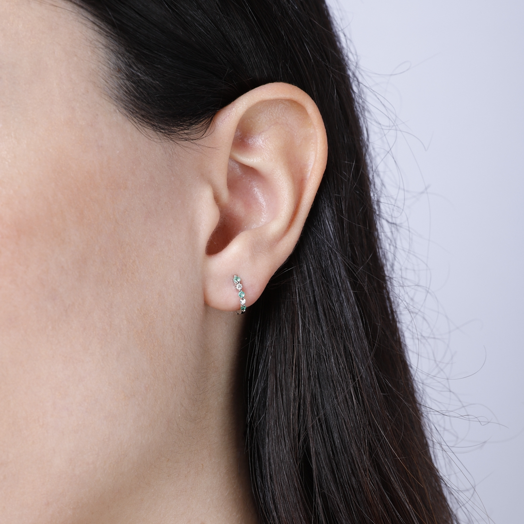 A woman with long brunette hair is wearing a pair of huggie earrings with alternating sapphire and white diamonds. 