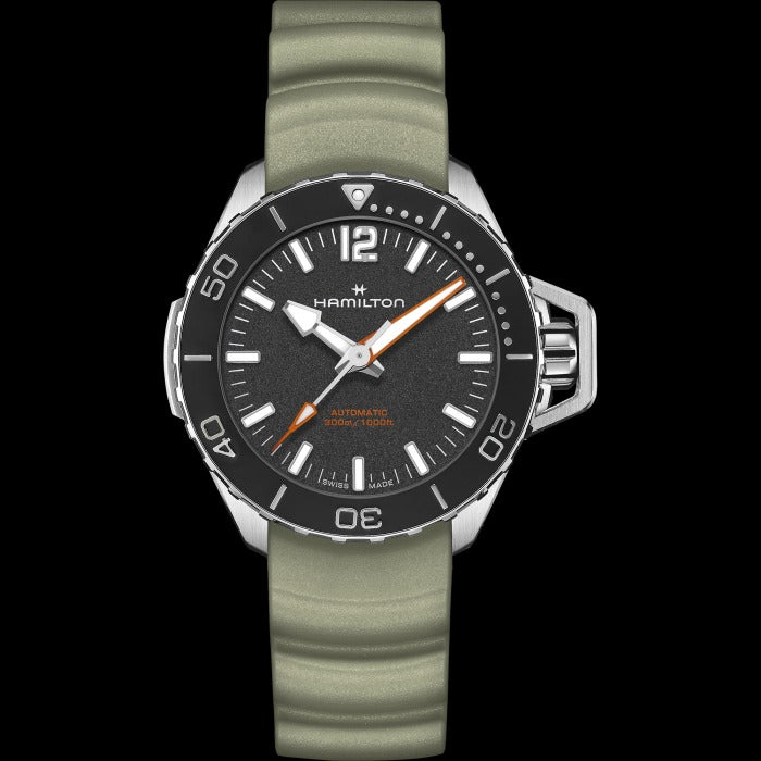 A Hamilton watch is laid out against a black background. It features a black dial, white hands and markers, a steel case, a black bezel, a steel crown, and a green rubber strap.