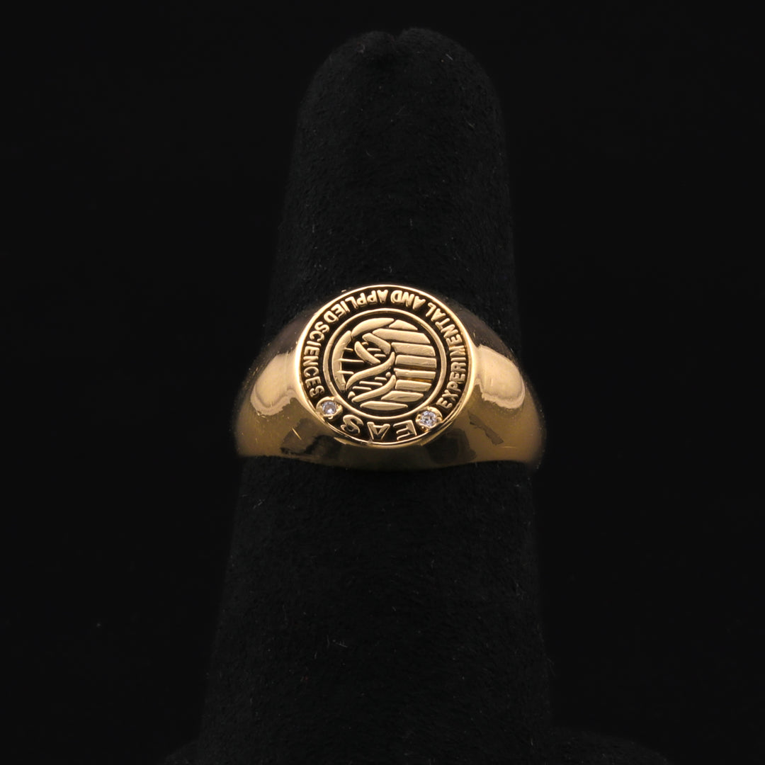 A close-up of a yellow-gold ring with an "Applied Sciences" stamped design and diamonds resting on a black ring holder against a black background. 