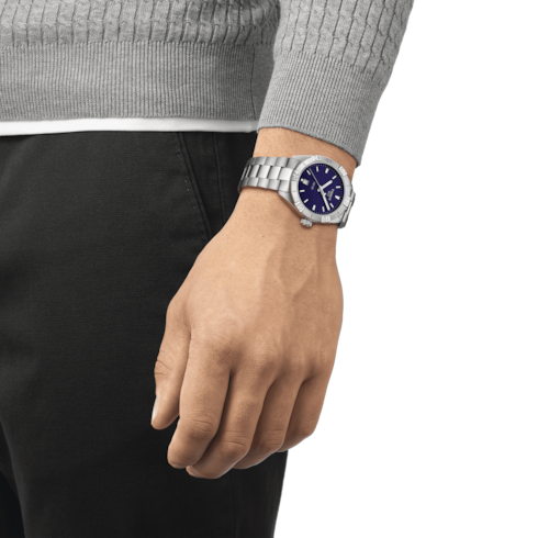 A woman wears a gray sweater, and a Tissot watch around her wrist. This is a close-up of a Tissot watch against a transparent background. The watch features a blue dial, a steel bezel, silver hands and markers, and a stainless steel bracelet.