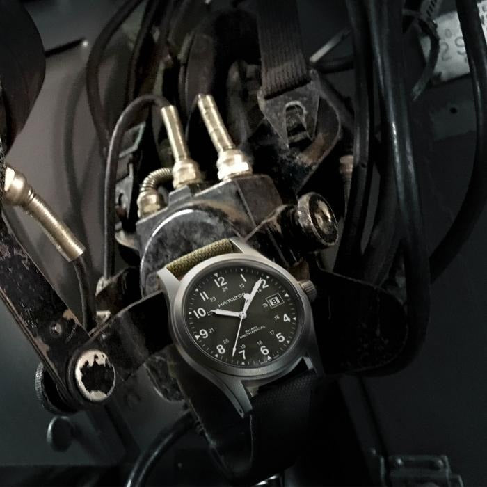 A Hamilton watch hooked between airplane machinery. It features a green dial, white hands and markers, a stainless steel case, a steel bezel, a steel crown, and a green canvas strap.