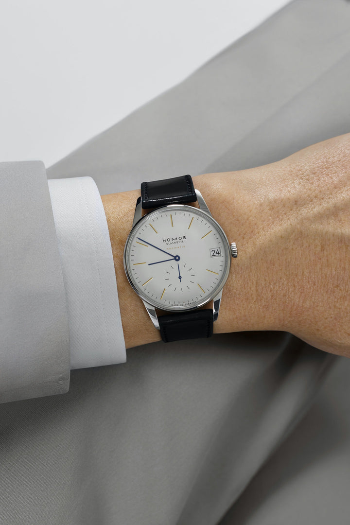 A man wearing a Nomos Glashutte watch on his wrist. It features a white dial, gold & blue hands and markers, a stainless steel bezel, and a black strap.