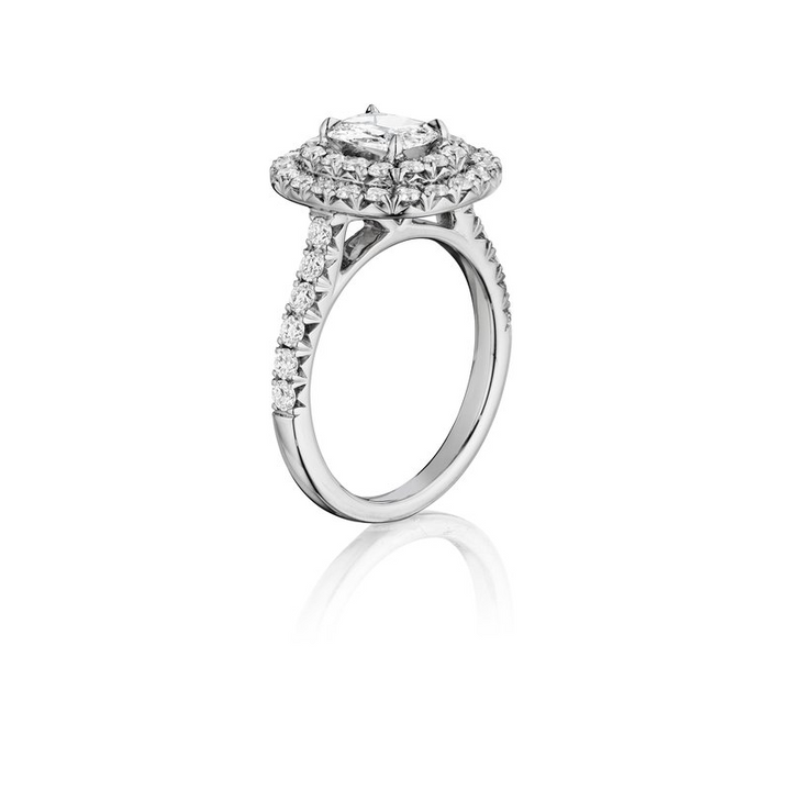 The Double Halo Engagement Ring, made of white gold. It is angled against a white background to the right, showing a side view of the ring. It features a brilliant-cut diamond set in the center, a double halo, and a round diamond-set band.