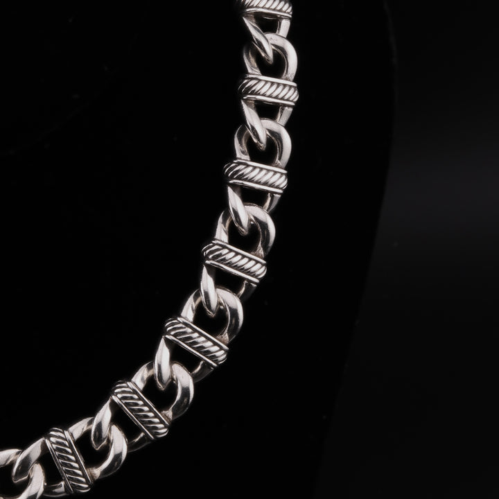 A close-up showing the right side of a sterling silver chain necklace with curb links and detailed stations against a black background.