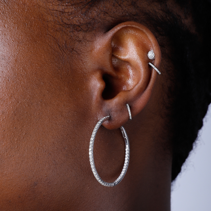 A woman with black short hair is wearing a pair of hoop earrings with a single row of round diamonds.