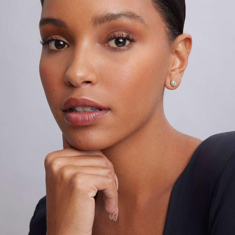 A woman is wearing a black top and a pair of two-toned stud earrings with a Pavé diamond circle motif framed by 18K gold Caviar beading. 