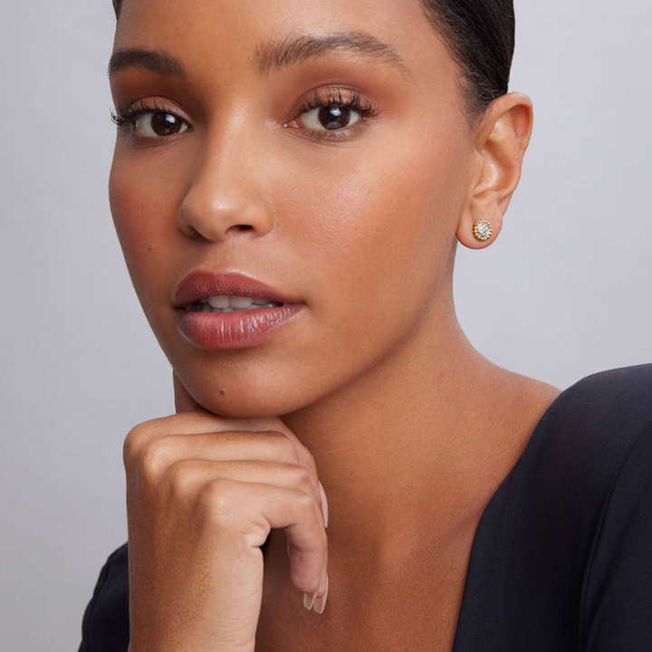 A woman is wearing a black top and a pair of two-toned stud earrings with a Pavé diamond circle motif framed by 18K gold Caviar beading. 