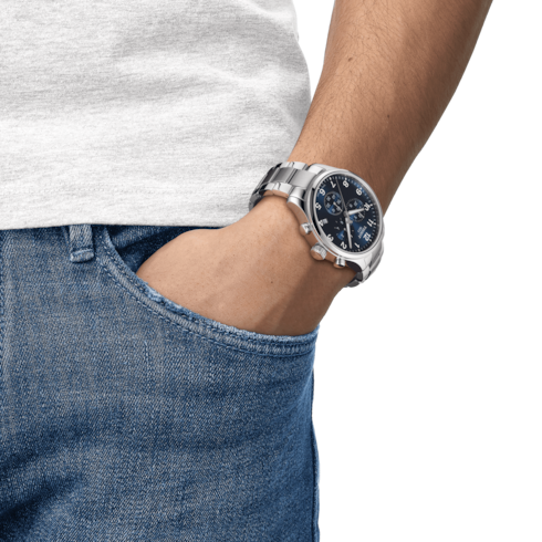 A man is wearing a Tissot watch around his wrist. The watch features a blue dial, white hands, markers, and stainless steel bracelet.