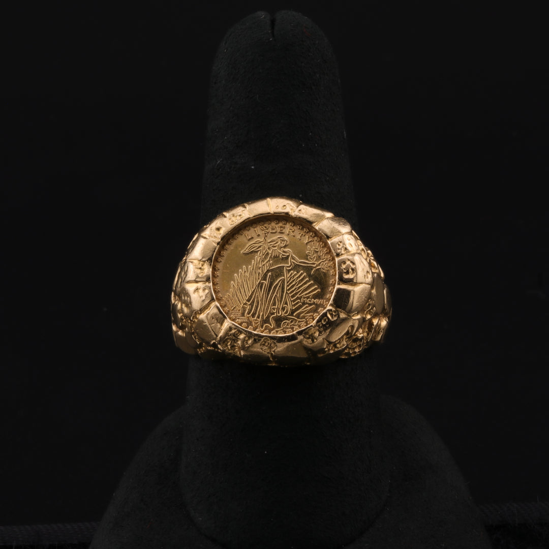 A close-up of a yellow-gold nugget ring with an authentic liberty stamp, set within a textured yellow gold design resting on a black ring holder against a black background. 